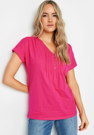 Lyserød kortærmet T-shirt i blødt stof, med V-hals, frontfolder og dekorative knapper. Sammen med blå jeans.