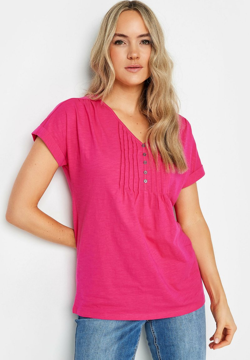 T-shirt rosa a maniche corte in un tessuto morbido, con scollatura a V, pieghe frontali e bottoni decorativi. Abbinato a jeans blu.