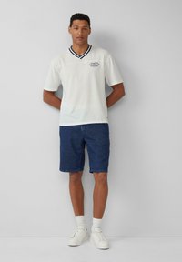 Witte polo met korte mouwen en marineblauwe accenten, gecombineerd met donkere denim shorts. Model staat met handen achter de rug, en draagt witte sneakers en sokken.