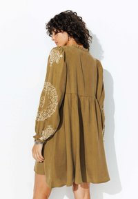 Robe vert olive en tissu doux, avec de longues manches bouffantes ornées de motifs brodés blancs, et une taille froncée pour une silhouette fluide.