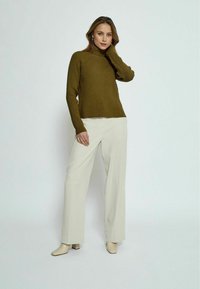 Olive zeleno rebrasto pulover s turtleneckom, v kombinaciji s svetlim bež širokimi hlačami in bež čevlji z visokim petakom, na svetli ozadju.
