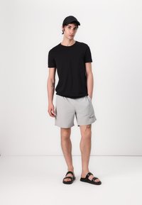 Tricou negru cu mânecă scurtă, pantaloni scurți gri deschis cu logo, sandale negre. Modelul stă pe un fundal neutru, evidențiind simplitatea ținutei.