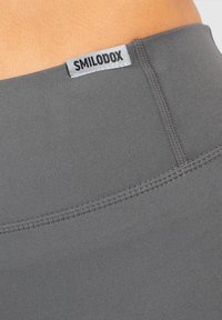Acercamiento de una tela elástica gris con costuras visibles y una etiqueta blanca que dice "SMILODOX" en la cinturilla.