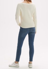 Pull crème en tricot avec un col large, associé à un jean bleu ajusté et des baskets blanches avec des accents verts.