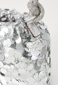 Silver sequin bucket bag made from shiny, reflective material. Features a knotted silver strap and a small rectangular tag.