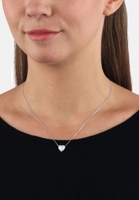 Collana con pendente a forma di cuore in argento su una catena sottile, caratterizzata da una superficie texture e un leggero luccichio. Indossata sopra un top nero a coste.
