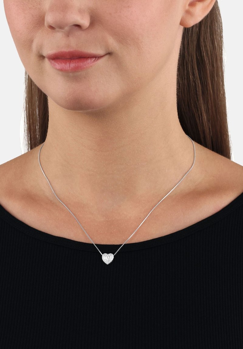 Collana con pendente a forma di cuore in argento su una catena sottile, caratterizzata da una superficie texture e un leggero luccichio. Indossata sopra un top nero a coste.