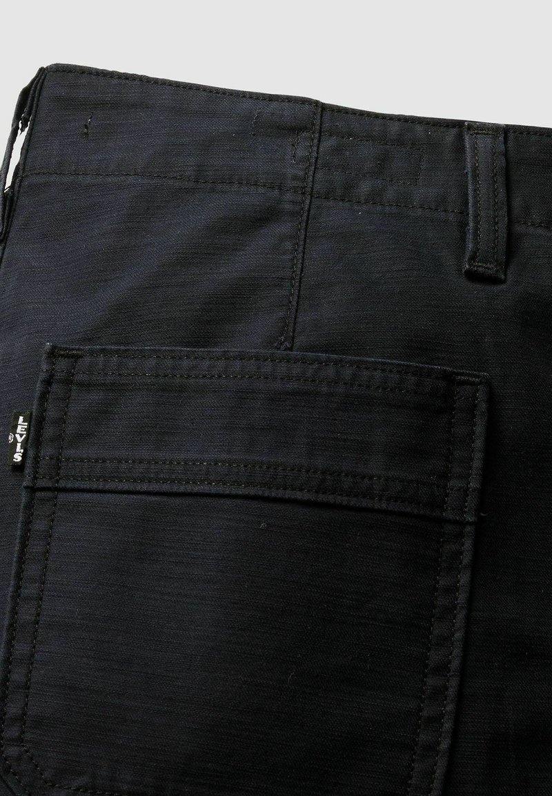 I pantaloni in denim nero presentano una tasca posteriore con cuciture visibili e un piccolo etichetta di marca. Il tessuto ha una texture liscia e una leggera lucentezza.