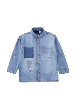Camicia di denim azzurro chiaro con chiusura frontale a bottoni, due tasche sul petto—una con toppa in tessuto più scuro—e tre toppe cucite sulla parte inferiore sinistra.