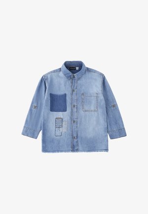 Camicia di denim azzurro chiaro con chiusura frontale a bottoni, due tasche sul petto—una con toppa in tessuto più scuro—e tre toppe cucite sulla parte inferiore sinistra.