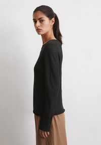 Zwarte lange mouwen top met een ronde halslijn en een gladde textuur, gecombineerd met een lichtbruine rok, getoond vanuit een zijprofiel.