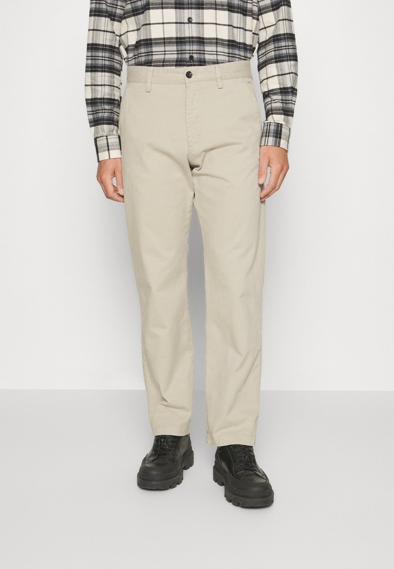 Mężczyzna ubrany w beżowe spodnie o prostych nogawkach, czarne masywne sznurowane buty i czarno-białą koszulę w kratę z długim rękawem.