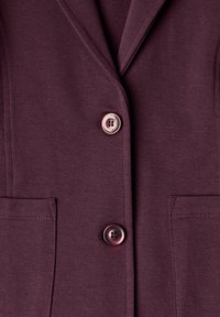 Gros plan d’une veste bordeaux avec deux boutons et deux poches avant, montrant le col et les détails de couture.