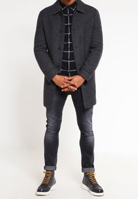 Uomo che indossa un cappotto lungo grigio scuro, una camicia a quadri bianco e nera, jeans skinny neri sbiaditi e stivaletti alti neri con lacci beige.
