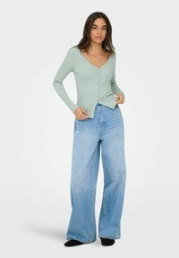 Jasnozielona prążkowana koszula na guziki z długim rękawem i dekoltem w serek, zestawiona z wysokim stanem i szerokimi nogawkami niebieskich dżinsów. Widoczne czarne skarpetki.