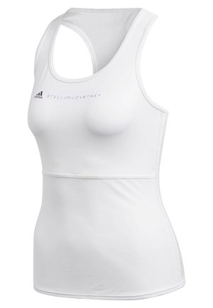 Femme athlétique portant un débardeur blanc et des leggings noirs avec des panneaux en mesh, debout, les bras relâchés le long du corps.