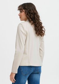 Beige skräddarsydd blazer med långa ärmar, strukturerade axlar och rena linjer, kombinerad med blå denimjeans. Slät tygtextur.