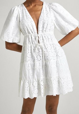 Kvinde iført en hvid mini nederdel i øjletsblonde med puffede korte ærmer, dyb V-udskæring, snørret talje og lagdelt nederdel.