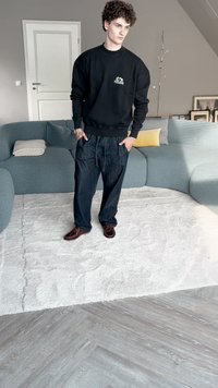Sweatshirt preto com logótipo, calças de ganga largas e sapatos castanhos-escuros. O indivíduo está de pé sobre um tapete branco texturizado numa sala moderna.