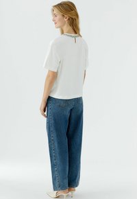 Weißes kurzärmeliges Oberteil mit einer Perlenbesetzung am Ausschnitt und einem Schlüssellochdetail am Rücken, kombiniert mit locker sitzenden blauen Jeans.