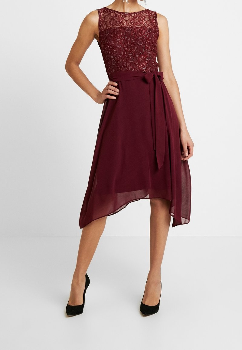 Ärmlös burgundy klänning med spetsliv, knytbälte i midjan och asymmetrisk chiffongsärk, bärs med svarta högklackade skor på en modell.