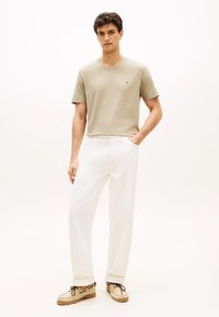 Μπεζ T-shirt με V-λαιμόκοψη, λευκό παντελόνι με ίσια γραμμή και μπεζ παπούτσια με καφέ κορδόνια. Ελάχιστο λογότυπο στο πουκάμισο, απλό σχέδιο.