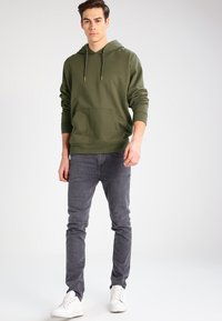 Felpa con cappuccio verde oliva con tasca frontale, abbinata a jeans slim-fit grigi e sneakers bianche. Tessuto morbido e design semplice.
