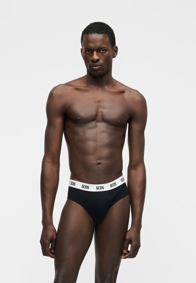 Homme athlétique debout devant un fond uni, portant un slip noir avec une ceinture blanche portant le logo "GCDS" répété.