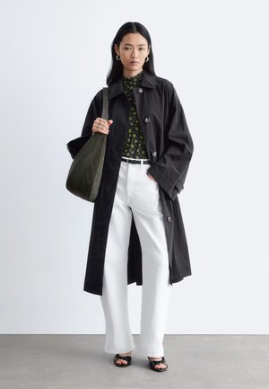 Kvinde iført en sort lang frakke, blomsterbluse, hvide bukser, sorte sandaler og bærende en stor mørkegrøn skuldertaske.