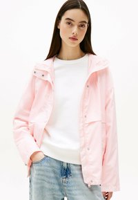 Giacca impermeabile rosa chiaro con cappuccio, chiusura a pressione e due tasche anteriori, abbinata a un maglione bianco e jeans azzurri chiari.