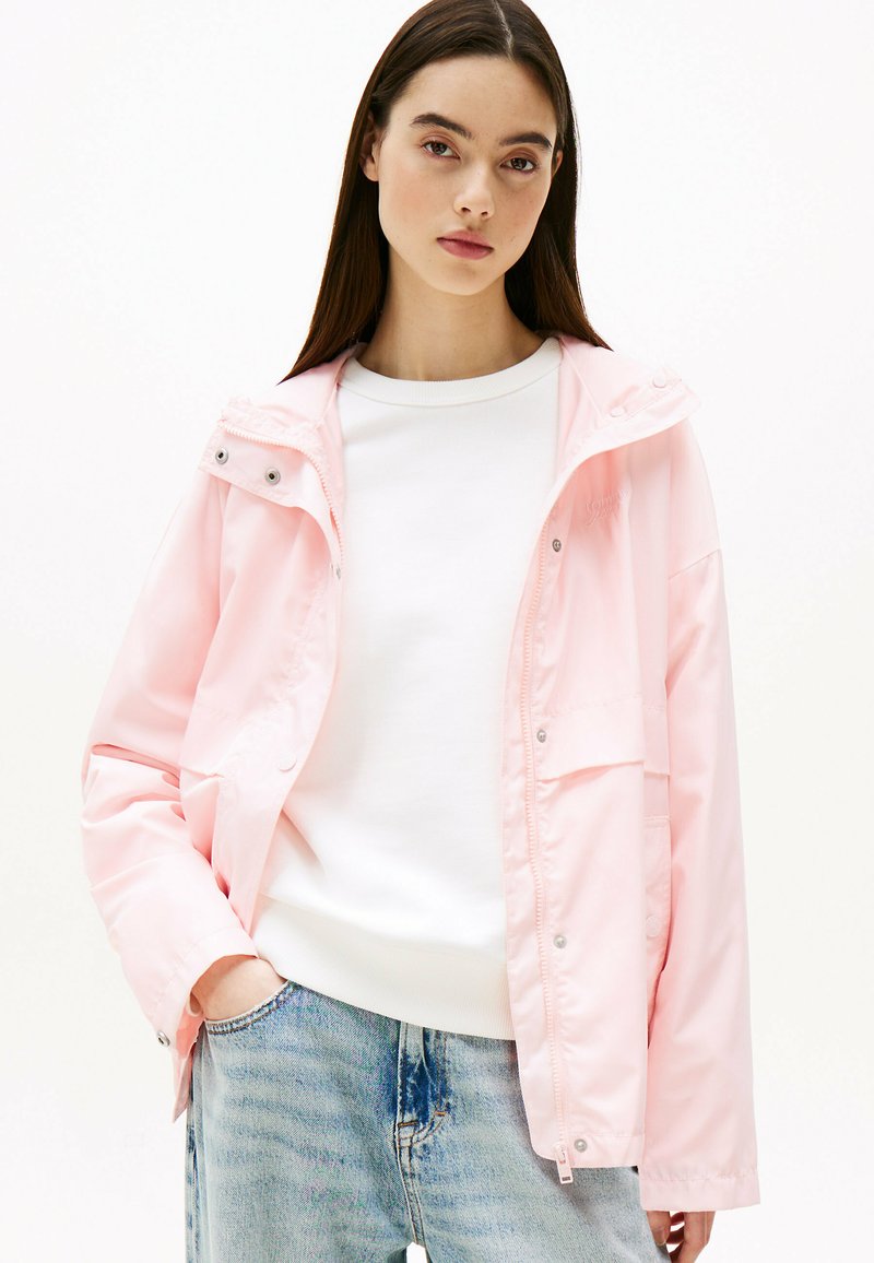 Giacca impermeabile rosa chiaro con cappuccio, chiusura a pressione e due tasche anteriori, abbinata a un maglione bianco e jeans azzurri chiari.