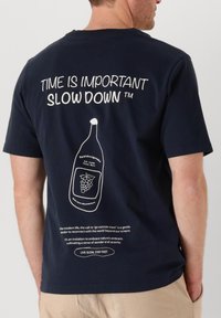 T-shirt in cotone blu con testo e grafica bianchi sul retro, incluso un'illustrazione di una bottiglia e uno slogan dettagliato. Maniche corte, scollo rotondo.