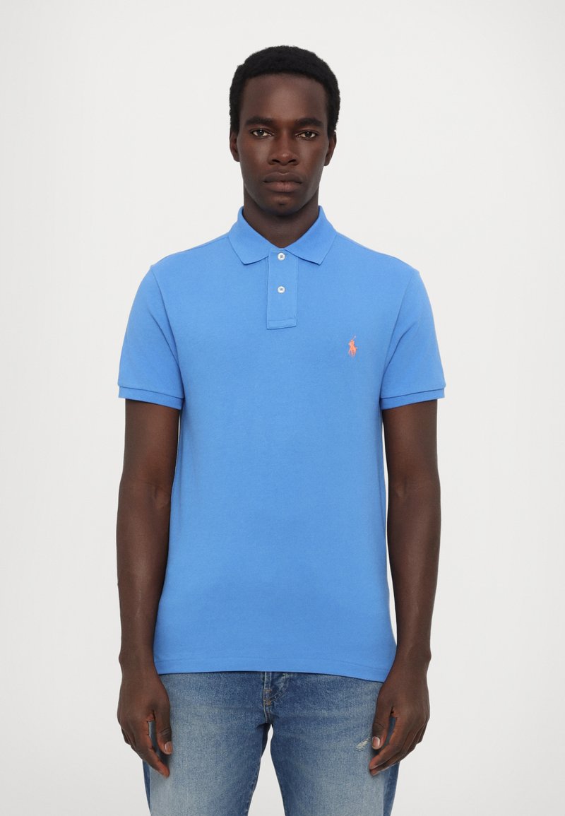 Zils kokvilnas polo krekls ar klasisku apkakli, trim pogām un mazu oranžu logo kreisajā krūtī. Nēsāts ar izbalējušām džinsu biksēm.