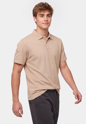 Ung mand med lyst brunt hår iført en beige kortærmet polo og mørke afslappede bukser, stående mod en ensfarvet baggrund.
