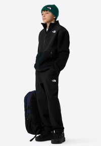 Schwarze Fleecejacke mit Reißverschluss, schwarze Hose, grüne Strickmütze und schwarze Wanderschuhe. Der Rucksack hat blaue und schwarze Akzente.