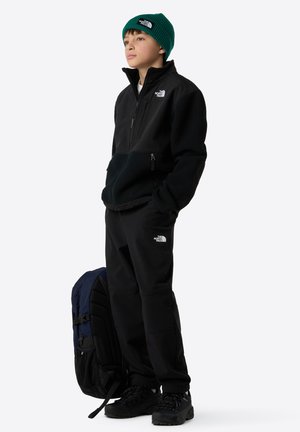 Giacca in pile nera con zip, pantaloni neri, berretto in maglia verde e scarponi da trekking neri. Zaino con accenti blu e neri.