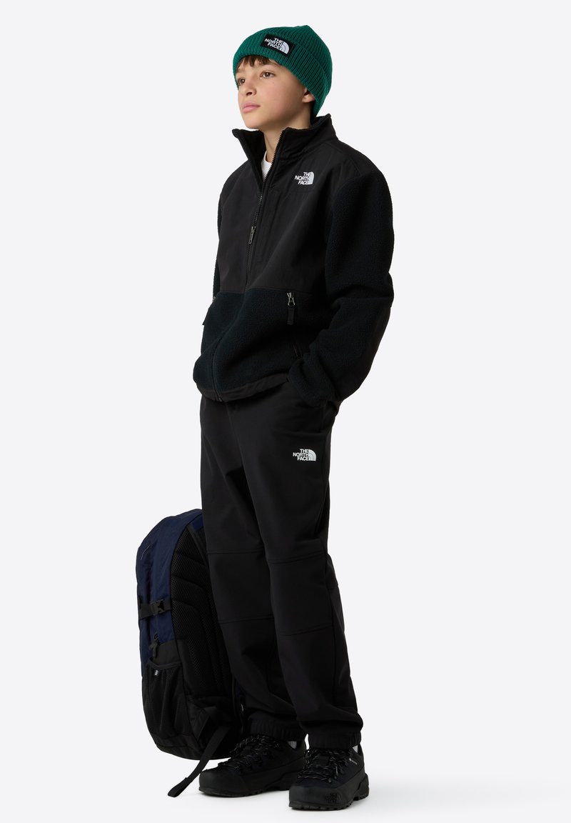 Schwarze Fleecejacke mit Reißverschluss, schwarze Hose, grüne Strickmütze und schwarze Wanderschuhe. Der Rucksack hat blaue und schwarze Akzente.