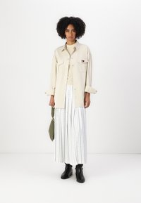 Beige button-up skjorte med to brystlommer, parret med hvite stripete vide bukser med fargerike prikker, og svarte ankelstøvletter.