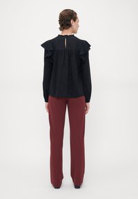 Blusa nera con maniche a volant, tessuto testurizzato, dettaglio a chiave sulla schiena, abbinata a pantaloni larghi color borgogna.