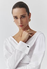 Witte blouse met lange mouwen en een diepe V-hals, met een zwarte knoop op de manchette. Gouden ronde oorbellen benadrukken de look. Glad textiel.