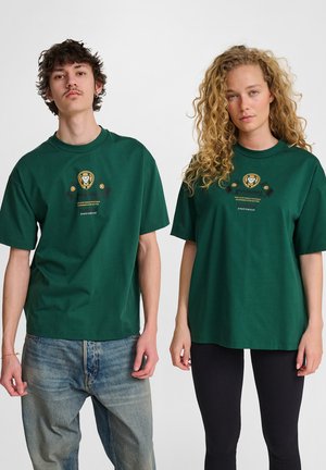 Deux modèles sont côte à côte portant des t-shirts graphiques vert foncé assortis ; le modèle de gauche porte un jean bleu, celui de droite porte des leggings noirs.