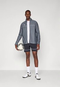 Gray athletic jacket with white accents, black shorts, white socks, and soccer ball. Model stands in neutral background.