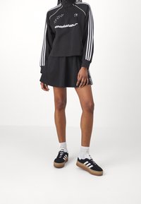Svart kort sweatshirt med vitt Adidas-logotyp och stjärndesign, parat med en svart kjol och svarta sneakers med vita ränder. Vita strumpor.