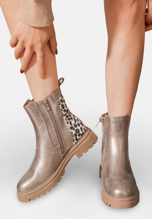 Bottines argentées métalliques avec une finition texturée, panneau latéral en imprimé léopard et semelle extérieure robuste à crampons. Fermeture zippée sur le côté pour un enfilage facile.