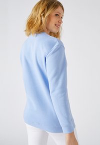 Sweatshirt azzurro chiaro con maniche lunghe, scollatura a girocollo e una texture di tessuto liscia. Anche dei pantaloni bianchi sono visibili nell'immagine.