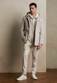 Jasnobeżowa kurtka z zamkiem, nałożona na beżowy hoodie i pasujące spodnie. Białe sneakersy dopełniają strój. Gładka tekstura, casualowy styl.