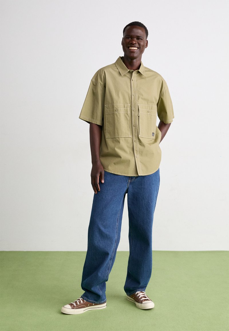 Camisa de manga corta verde oliva con dos bolsillos en la parte delantera, combinada con un pantalón vaquero azul holgado y zapatillas marrones. Diseño simple y funcional.