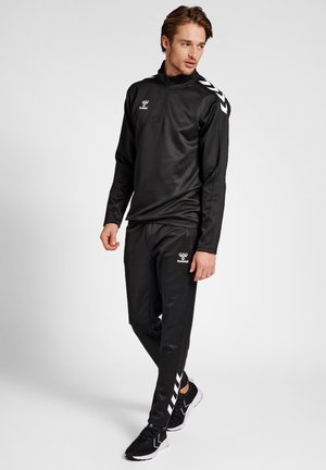 Zwarte sportieve trainingsoutfit met een ritskraag, voorzien van een wit logo en chevronaccenten op de mouwen en broek, gemaakt van soepel, lichtgewicht stof.