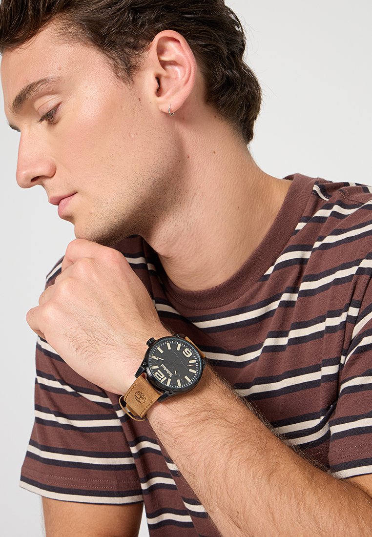 Un reloj negro con una carátula redonda y una correa de cuero beige. La correa presenta un logo, mientras que la carátula incluye números grandes y una textura sutil.