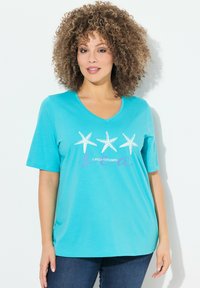 T-shirt turchese a maniche corte con scollatura a V, caratterizzato da tre stelle marine bianche e testo viola che recita "HO BISOGNO DI VITAMINA MARE". Tessuto di cotone morbido.